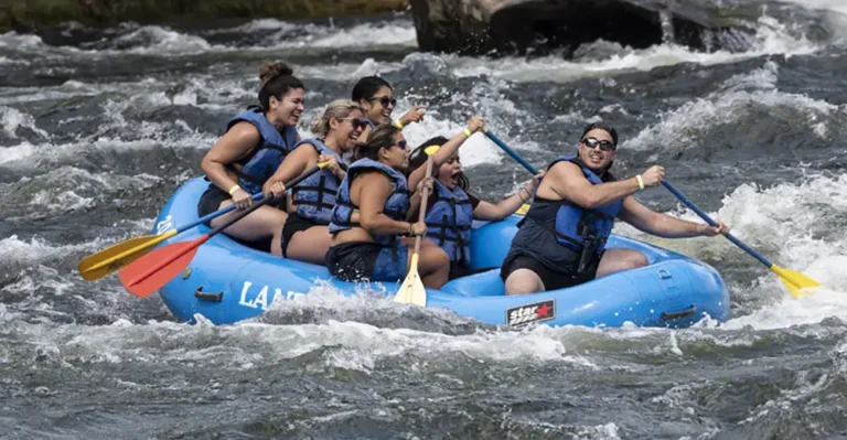 A group of six people in life jackets paddling a blue raft through white water rapids, enjoying an adventure on the river.