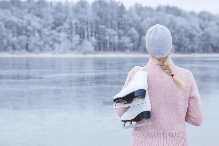 A person stands by an icy lake, holding ice skates, with a snowy forest backdrop.