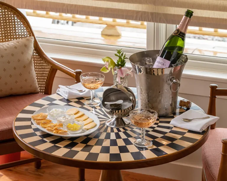 A cozy table setting featuring champagne, drinks, snacks, and fresh flowers, with a checkered table surface and natural light streaming in.
