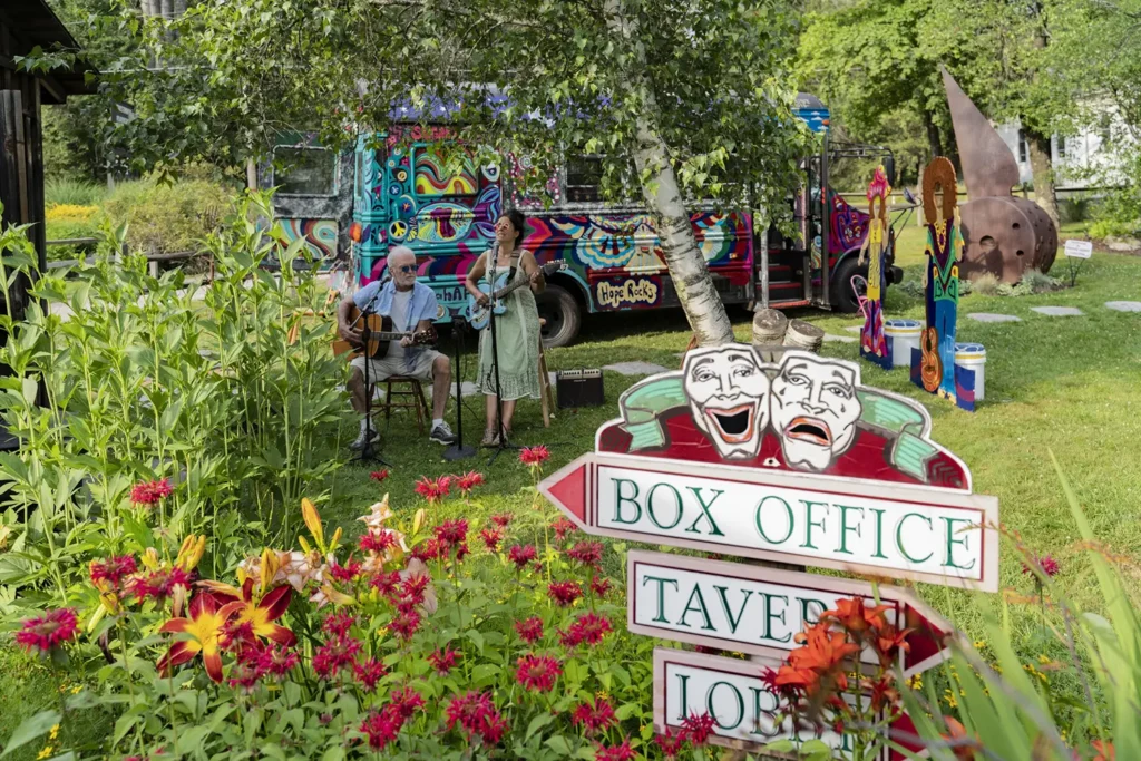 A colorful outdoor scene featuring musicians, vibrant flowers, and a signpost indicating various attractions.
