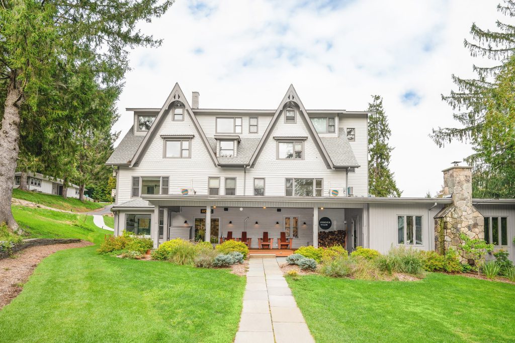 A large, white, three-story house with steep gabled roofs and a covered porch sits amid lush green lawns and tall trees under a partly cloudy sky—perfect for enjoying exclusive Catskills spring offers.