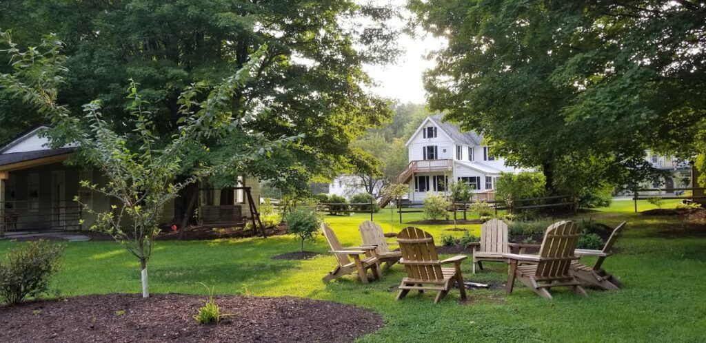 A grassy yard with several wooden Adirondack chairs arranged around a fire pit, surrounded by trees and bushes, with two white houses and porches in the background—perfect for relaxing and enjoying exclusive Catskills spring offers on a sunny day.