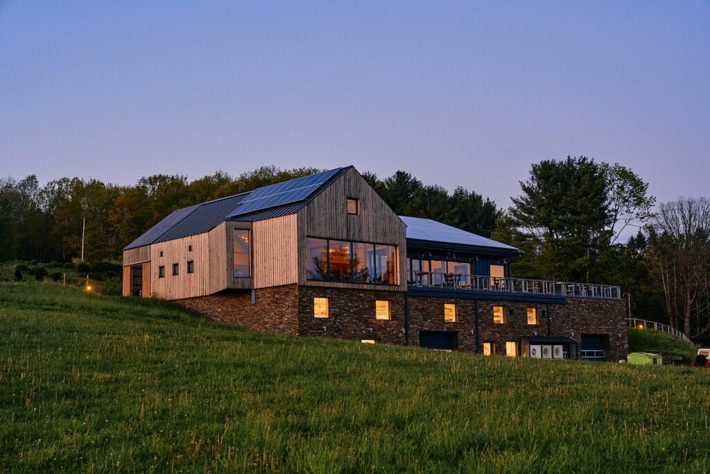 A modern, barn-style house with wood siding and large windows sits on a grassy hill at dusk. The interior lights glow warmly, and a stone foundation is visible beneath the house. Trees line the background.