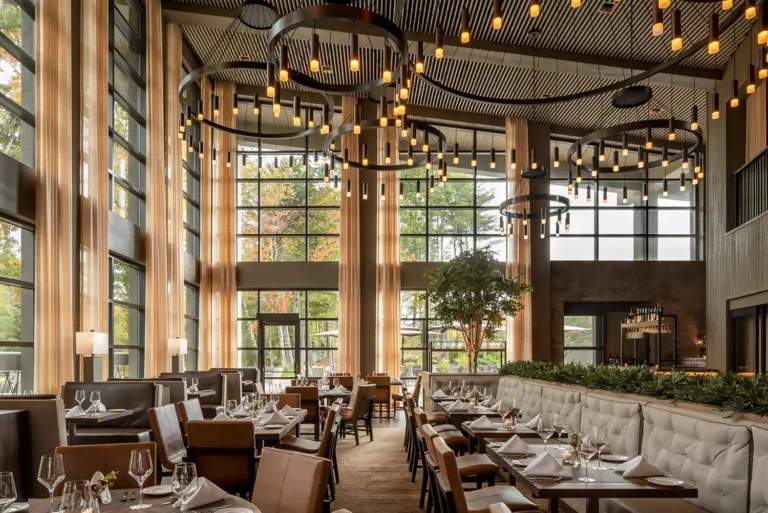 A modern restaurant interior featuring large windows, stylish lighting fixtures, and elegant seating, surrounded by greenery.