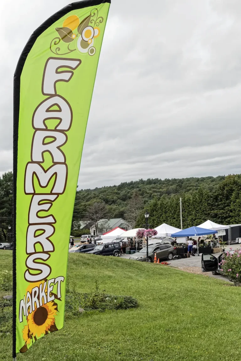 A vibrant green banner announces a farmers market, surrounded by tents and vehicles in a scenic outdoor setting.