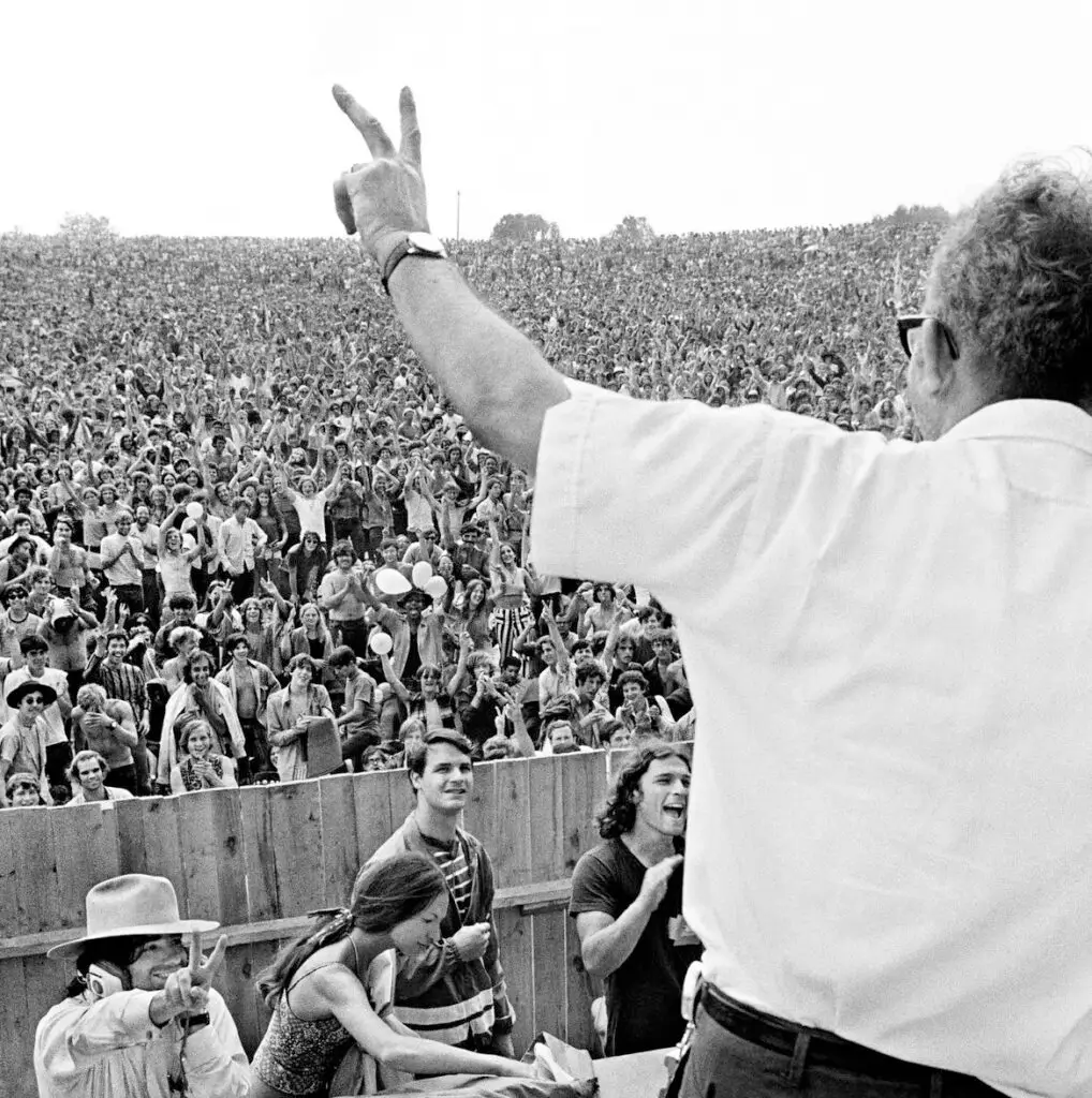 A large crowd gathered outdoors with a speaker raising a hand in a peace gesture, expressing enthusiasm and connection.