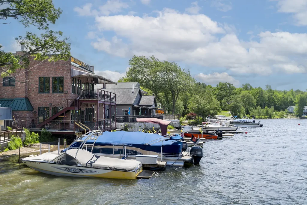 A tranquil lakeside scene with various boats docked alongside a tree-lined shore and a few quaint buildings in the background.