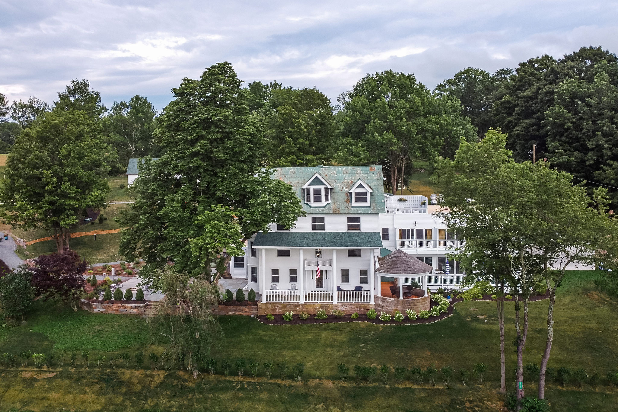 A large white two-story house with a green roof is surrounded by trees and a well-kept lawn. Landscaping and a gazebo are visible in the front yard under a cloudy sky.