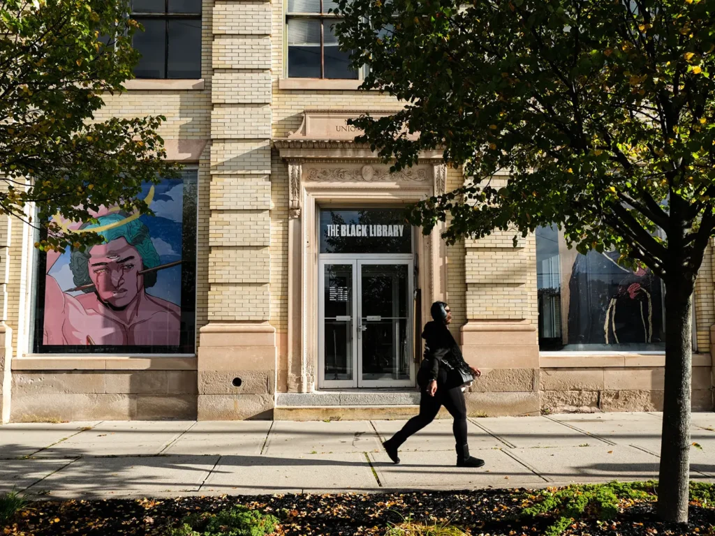 A person walking past a library's entrance with colorful artwork displayed in large windows, framed by trees along the sidewalk.