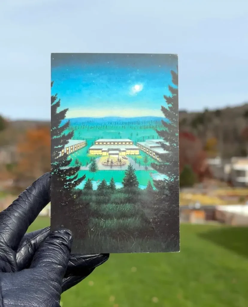 A hand holding a postcard with a serene landscape, featuring trees, a lake, and buildings under a clear blue sky.