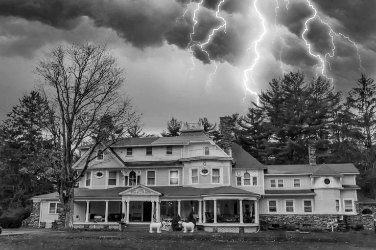 A large, vintage house under a stormy sky, illuminated by dramatic lightning, surrounded by tall trees in a black and white setting.