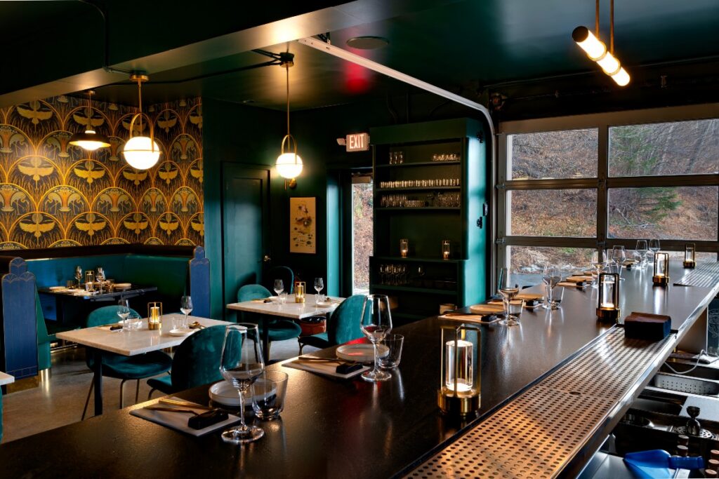 The stylish interior of Korean Threshold restaurant featuring a bar, teal chairs, and decorative walls, illuminated by modern lighting with a view through large windows.