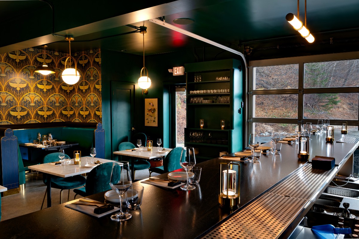 The stylish interior of Korean Threshold restaurant featuring a bar, teal chairs, and decorative walls, illuminated by modern lighting with a view through large windows.