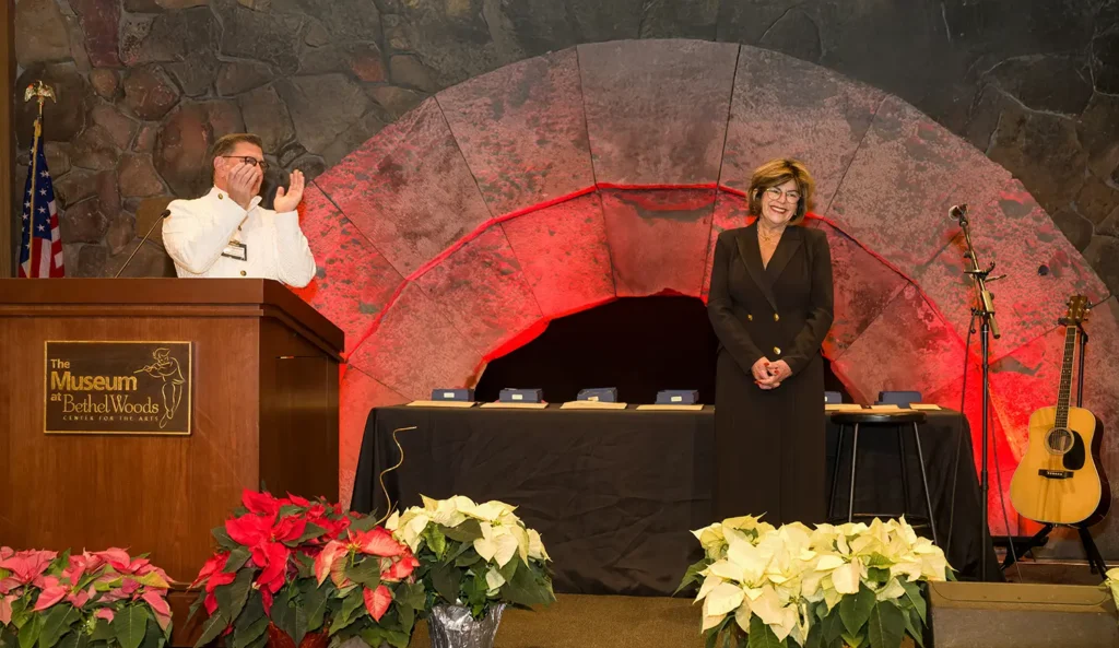 A formal event featuring a speaker and a recipient by a decorated stage, with festive decorations and music setup in the background.
