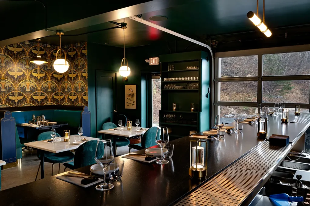 A stylish restaurant interior features dark green walls, modern lighting, and a bar area with elegant table settings.