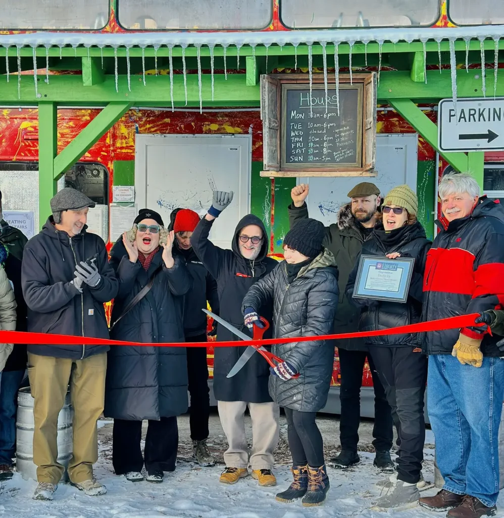 A group of people celebrate a ribbon-cutting ceremony outside a colorful building, holding signs and cheering amidst a snowy landscape.