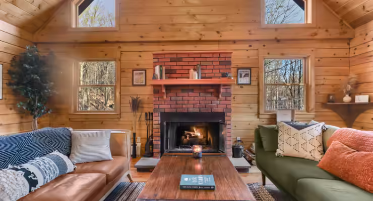 A cozy wooden cabin living room with a lit brick fireplace, two large sofas with patterned pillows, a wooden coffee table, and windows letting in natural light from the surrounding trees.