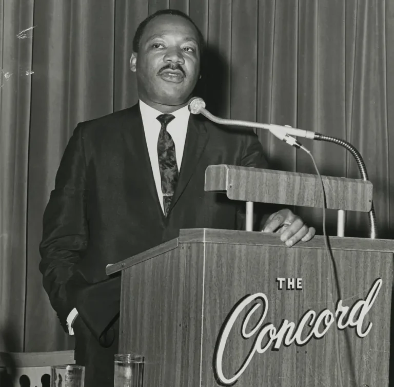 A man in a suit stands at a podium with a microphone, addressing an audience, with 'The Concord' visible on the front.