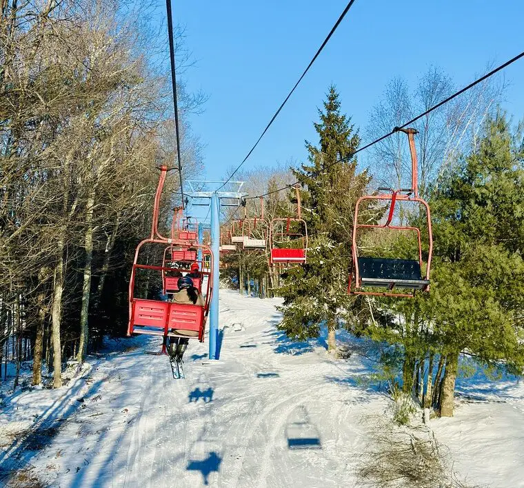 A ski lift with red chairs moving through a snowy landscape and tall trees under a clear blue sky.