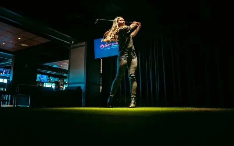 A woman swings a golf club in a dimly lit venue, showcasing her athletic posture against a dark background.