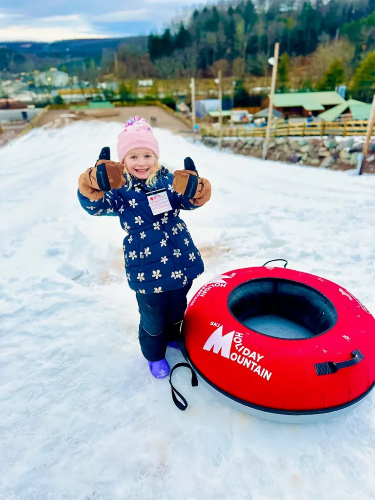 A happy child wearing winter clothing plays in the snow, holding up her hands with mittens near a red snow tube.