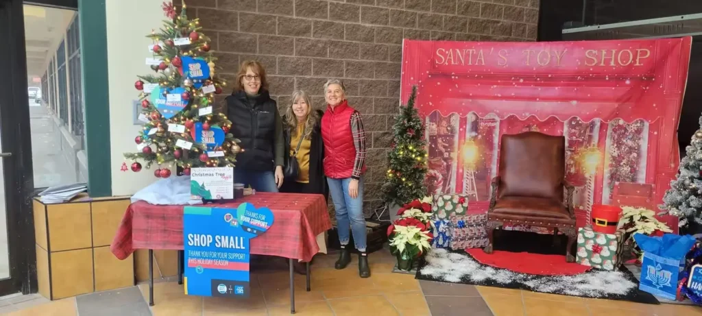 Three women stand at a holiday-themed booth with a decorated Christmas tree and a festive backdrop featuring a chair.