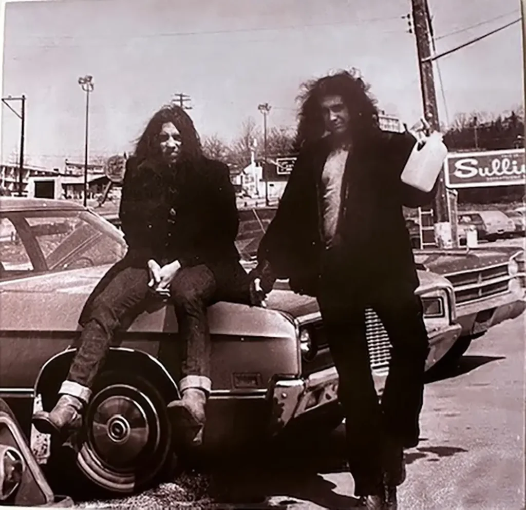 Two men with long hair stand by an old car, one holding a milk jug, set in a vintage, urban backdrop.