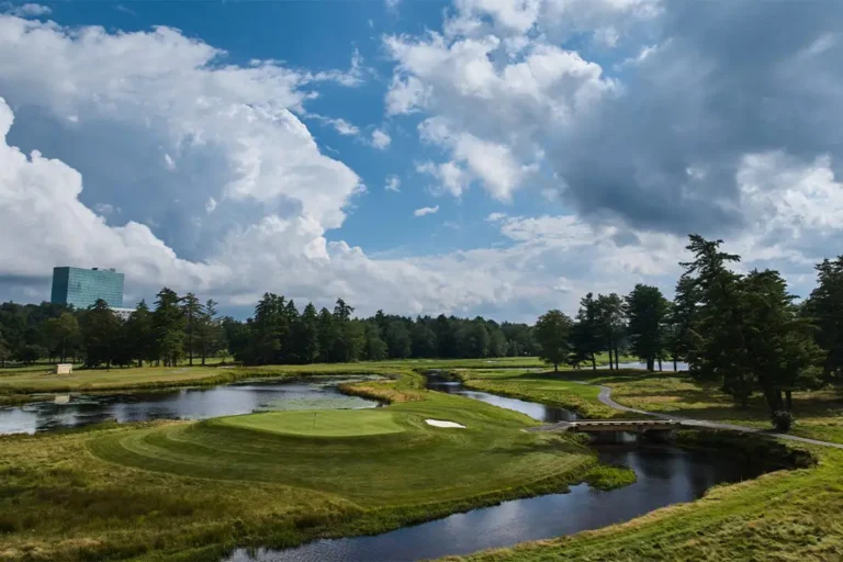 Lush green golf course with a winding stream, surrounded by trees and under a bright blue sky with fluffy clouds.