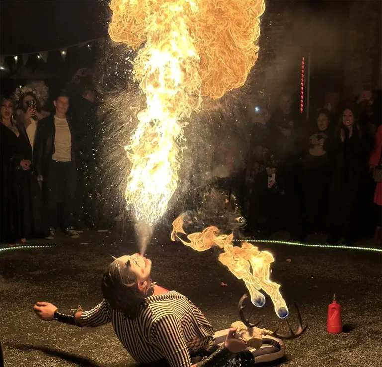 A performer lying on the ground breathes a large plume of fire, captivating an engaged audience at a lively outdoor event.
