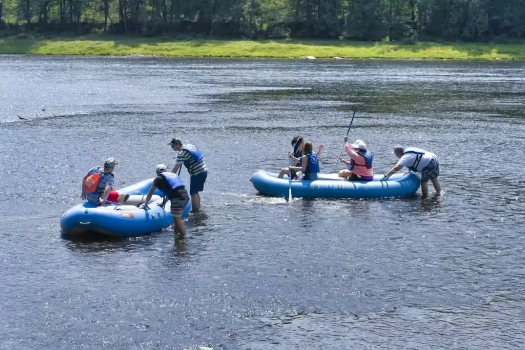 Several people are in rafts on a calm river, some paddling while others adjust their equipment, surrounded by greenery.