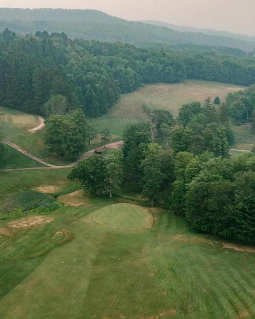 A scenic view of a green golf course surrounded by lush trees and rolling hills under a hazy sky.