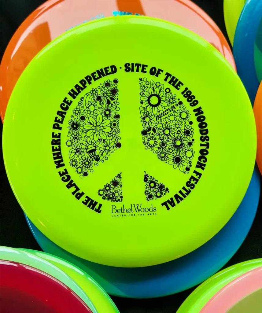 A vibrant green frisbee features a peace symbol and text referencing the 1969 Woodstock Festival and Bethel Woods.