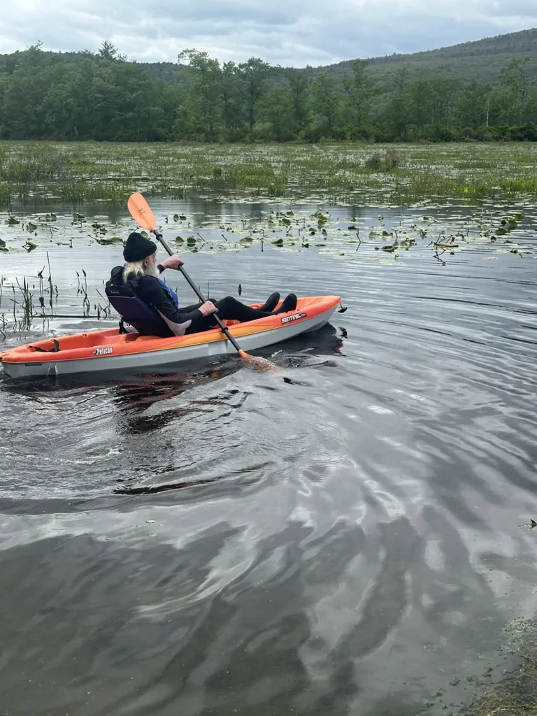 A person paddles a kayak through calm water surrounded by greenery and lily pads under a cloudy sky.