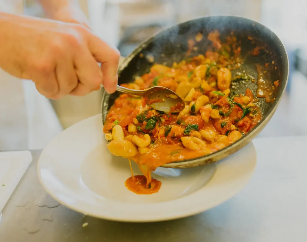 A chef serving a colorful pasta dish from a skillet onto a white plate, with steam rising from the food.
