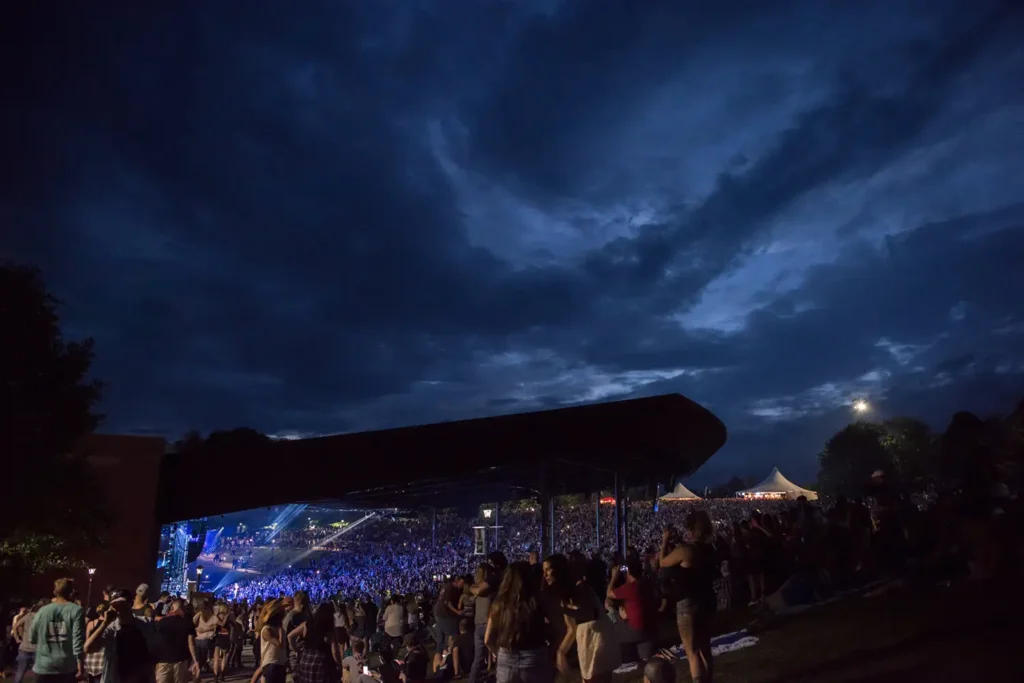A large crowd enjoys a concert under a dramatic, cloud-filled sky during twilight.