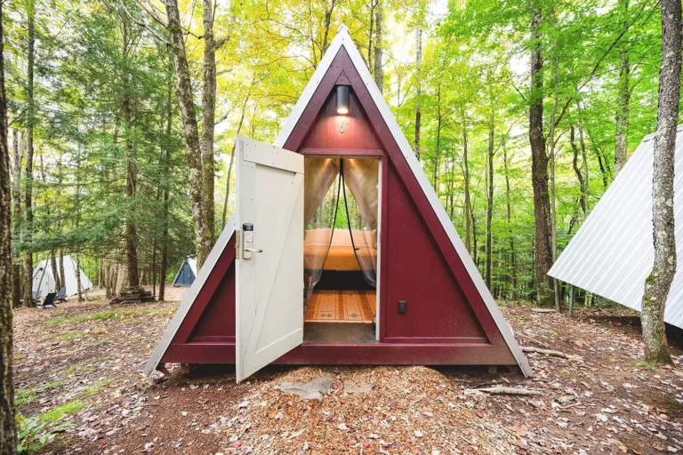 A triangular cabin nestled among trees, with an open door revealing interior wooden beams and a soft light overhead.