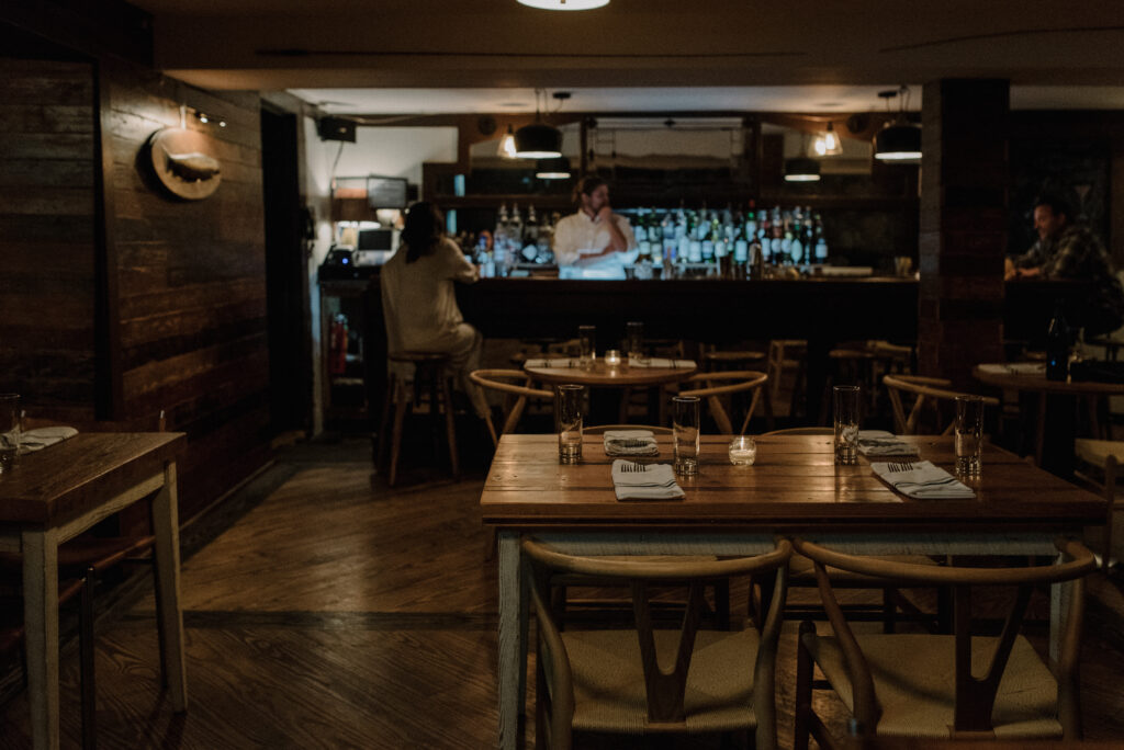 A dimly lit restaurant with wooden tables and chairs, set with napkins and glasses. A bar with bottles lines the back, where a bartender stands and two patrons sit on stools. The atmosphere is cozy and intimate.