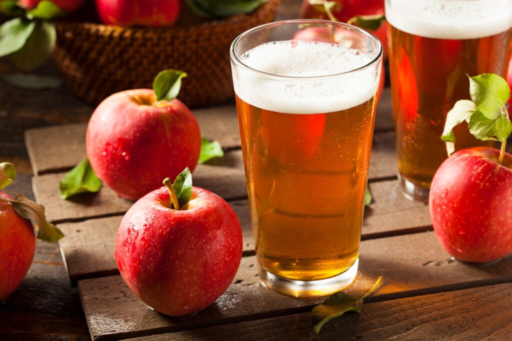 Two glasses of apple cider with foam on top sit on a wooden surface, surrounded by fresh red apples with green leaves. A basket of apples is visible in the background.