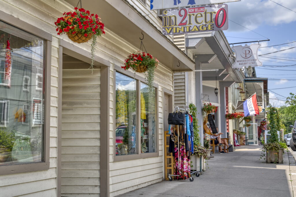 A sidewalk lined with storefronts, vintage and clothing shop signs, hanging flower baskets, and a rack of colorful clothes outside, on a sunny day in a small town.