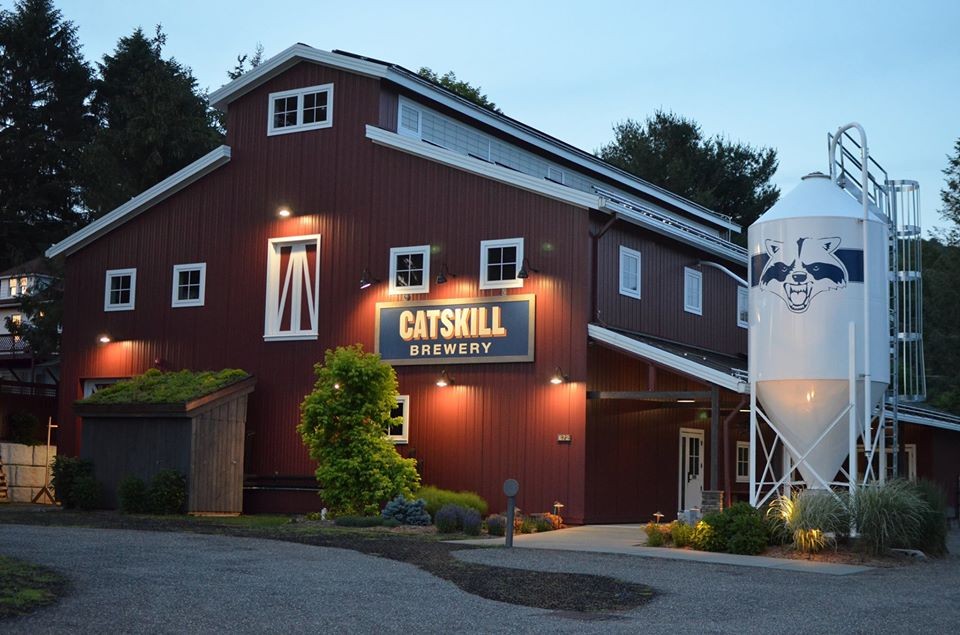 A red barn-style building with a lit Catskill Brewery sign on the front, surrounded by greenery, and a white silo with a cat face logo next to it, photographed at dusk.