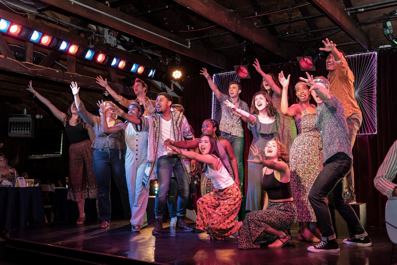 A diverse group of performers on stage, smiling and reaching forward with enthusiasm under colorful stage lights, in a lively theater setting with a wood-paneled ceiling and audience tables visible.