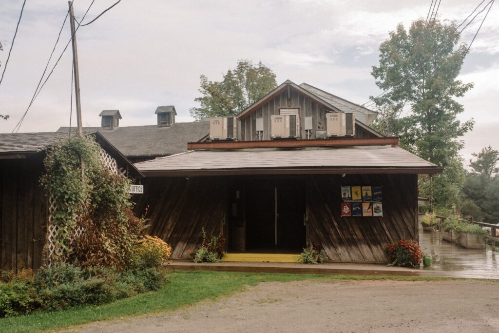 A rustic wooden building with a slanted roof, several air conditioning units on top, and colorful posters by the entrance, surrounded by greenery on a cloudy day.