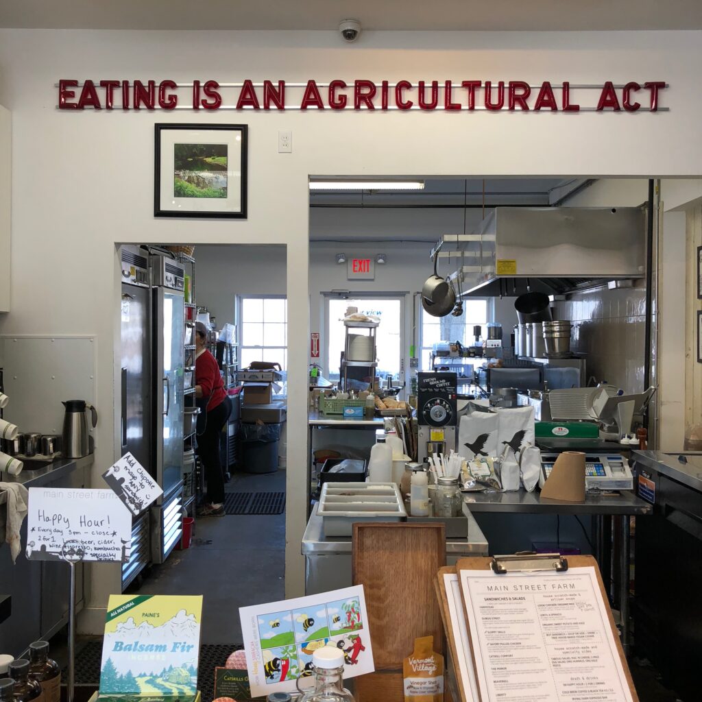 A café kitchen with cooking equipment and supplies, a person working in the back, and a red sign overhead reading “EATING IS AN AGRICULTURAL ACT.” Menus and a book display are visible in the front.