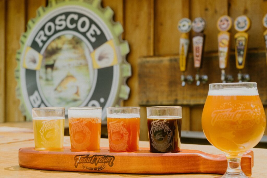 A wooden paddle holds four small beer glasses of different colors next to a full pint of beer. Behind them are beer taps and a large sign that reads Roscoe with a fish logo. The setting appears rustic and outdoors.