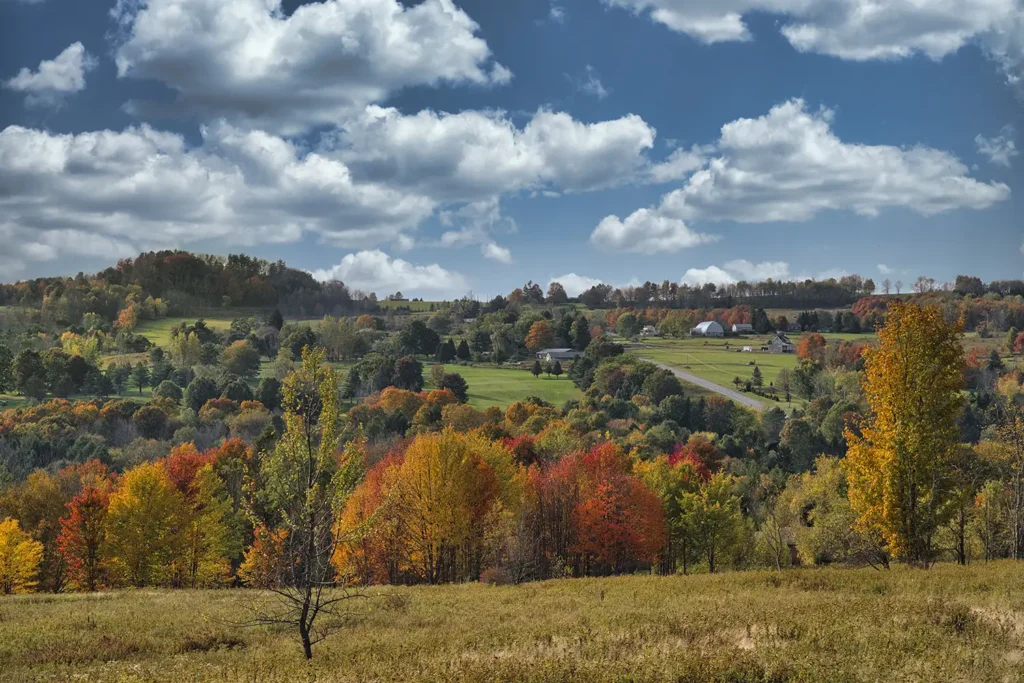 A vibrant autumn landscape with colorful trees, rolling hills, and a blue sky adorned with fluffy clouds.
