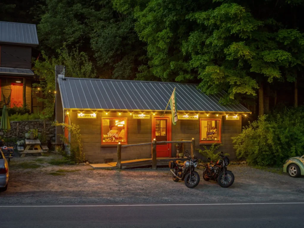A cozy café with warm yellow lights shines at dusk, surrounded by greenery. Two motorcycles are parked in front, and a ramp leads to the entrance. The windows display signs for coffee, sandwiches, and hand pies.