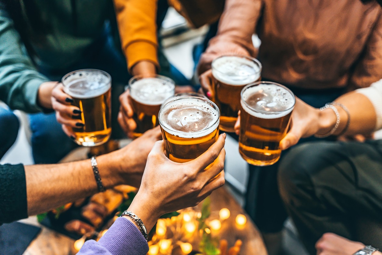 A group of people clinking glasses of beer together in a celebratory toast, with hands visible and blurred food and lights in the background.