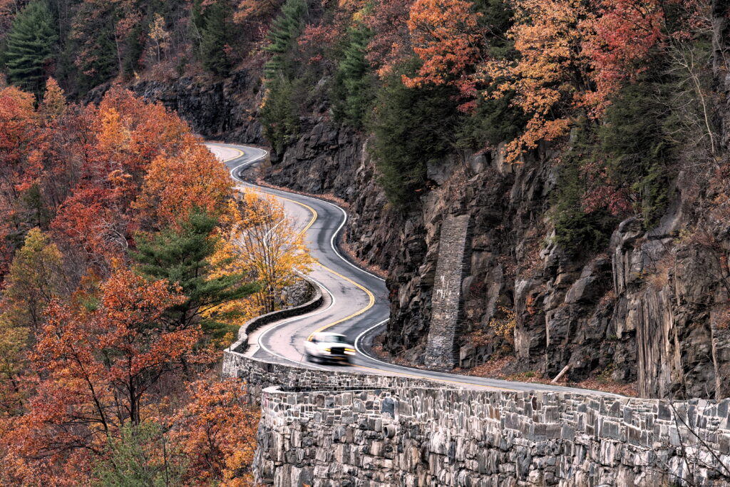 A winding road curves through rocky cliffs and vibrant autumn trees, with a blurred car driving along it. The scene is surrounded by red, orange, and yellow foliage.