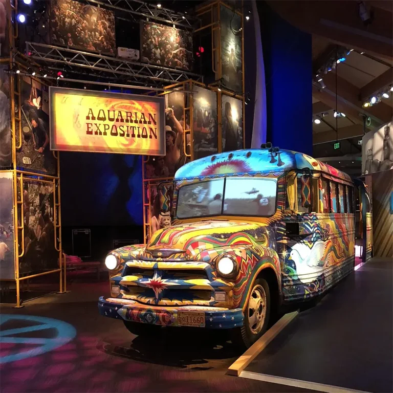 A colorful bus under vibrant lighting, positioned in an exhibition space with a sign that reads 'Aquarian Exposition' in the background.