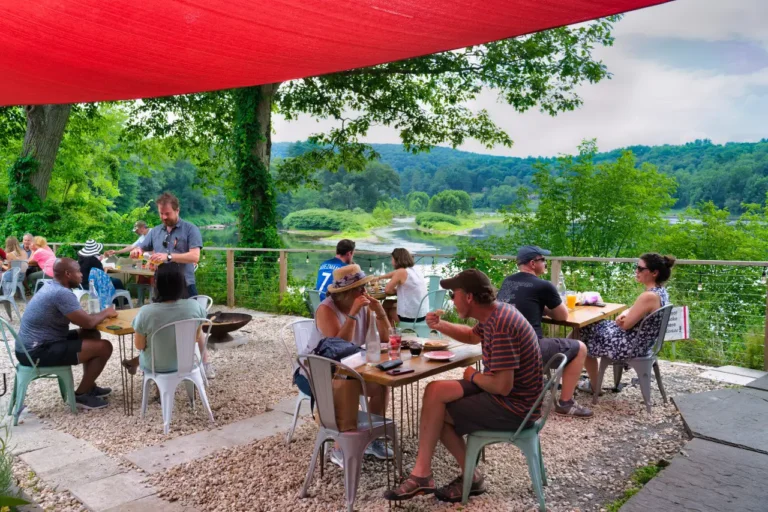 A lively outdoor dining scene with people enjoying meals under a red canopy, surrounded by greenery and a river view.